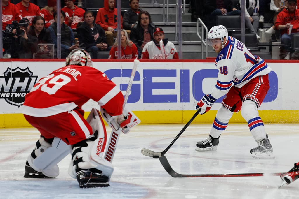 Player #18 in Rangers uniform skating with puck toward opposing goaltender in red jersey