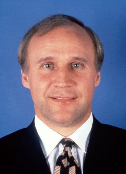 Professional headshot of a man in a dark suit and patterned tie against a blue background