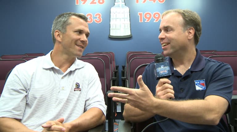Two men in polo shirts having a conversation with a microphone, seated in front of a backdrop