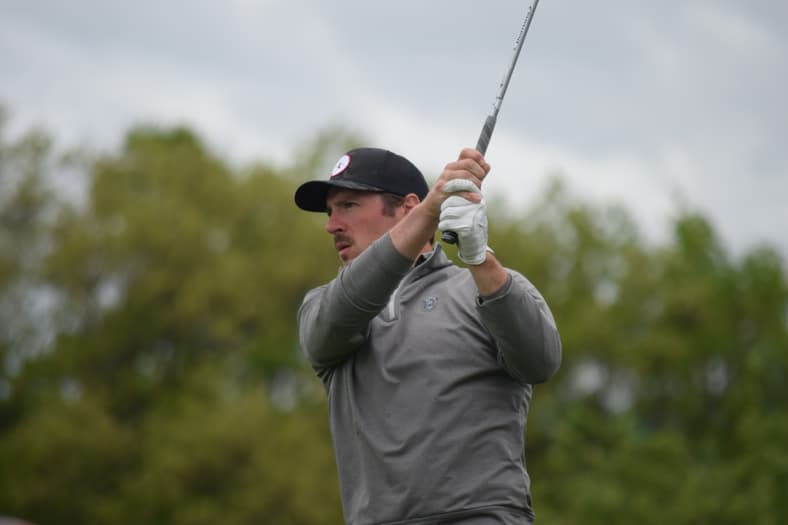 Golfer in gray shirt and black cap mid-swing on course with trees in background