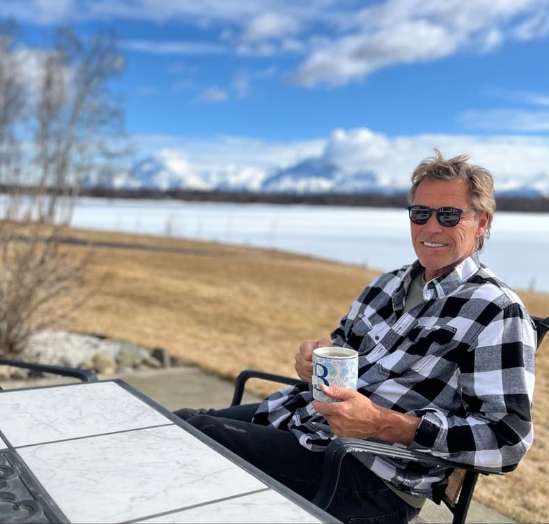 Man in plaid shirt holding mug overlooking snowy mountain lake landscape